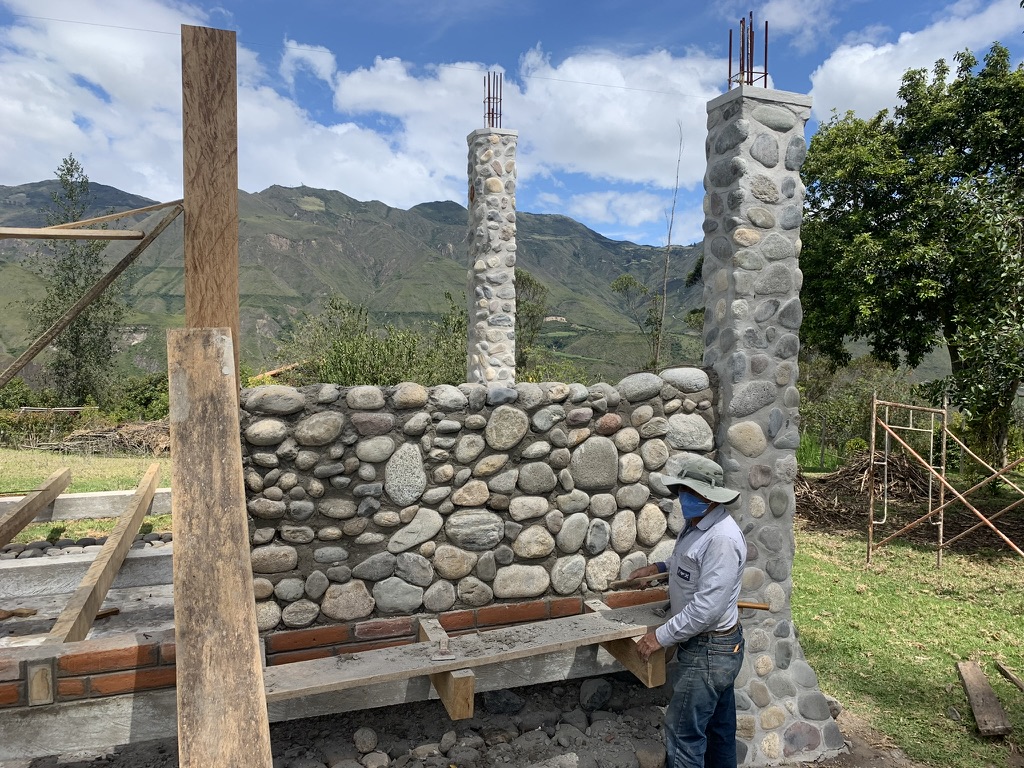 A mason in a sun hat working on a river-stone wall with mountains in the distance