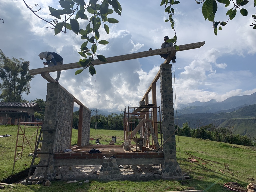 Two workers sitting atop a ridge beam being installed across the stone pillars